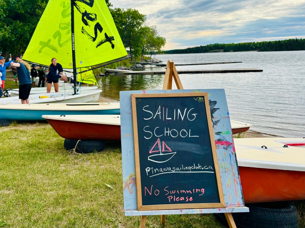 Sailing school sign written on a chalkboard with sailboats and a lake in the background